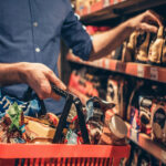 Mano tomando snacks del lineal con cesta llena, artículos típicos de compra por impulso en supermercado