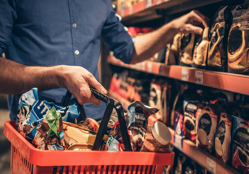 Mano tomando snacks del lineal con cesta llena, artículos típicos de compra por impulso en supermercado